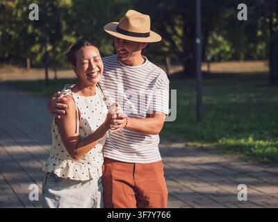 A happy young couple walks together through a peaceful park on a warm sunny day. They share a genuine laugh, capturing a relaxed moment filled with af Stock Photo
