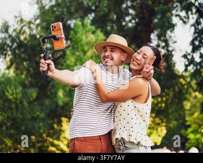 Joyful man and woman filming themselves together with a mobile phone stabilizer in a beautiful green park. Stock Photo