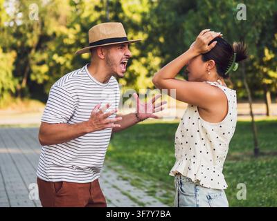 Young couple having an emotional argument outdoors, with the man expressing frustration while the woman holds her head in distress. Stock Photo