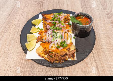 Close-up of birria-style quesatacos with cilantro, onion, lime wedges, and consommé on a slate plate. Stock Photo