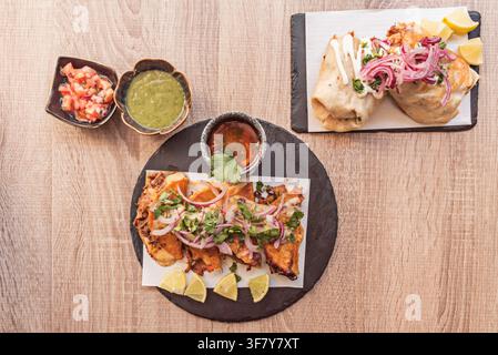 Overhead spread of birria quesatacos, burritos, consommé, pico de gallo, and salsa verde on wood table. Stock Photo