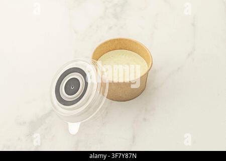 Herb-flecked creamy dipping sauce in open kraft paper cup with clear lid beside it on marble. Stock Photo
