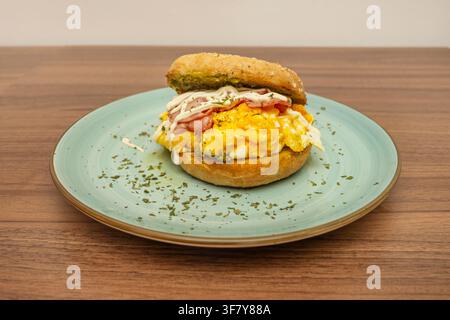 Breakfast sandwich with scrambled eggs, ham, and mayo drizzle on sesame bun, plated in teal dish. Stock Photo
