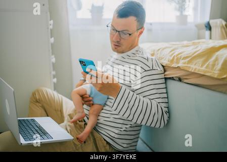 Father working from home, holding his baby while using a laptop and phone, representing the challenges and realities of modern parenting and professional life Stock Photo