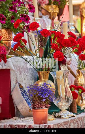Red carnations arranged in a brass vase form the focal point of a richly layered display with purple flowers, glassware, lace textiles and traditional Stock Photo