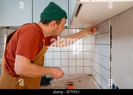 Man in work overalls and green beanie carefully applies grout spacers between white ceramic tiles on a kitchen wall, undertaking a renovation project and doing a home improvement task Stock Photo