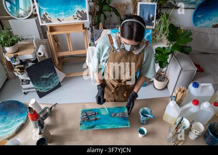Woman crafter in apron and protective respirator creating epoxy resin art at worktable in workshop. Stock Photo