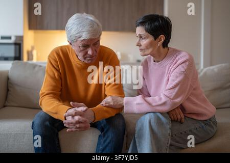Concerned attentive elderly wife supporting emotionally stressed husband sitting on sofa at home. Stock Photo