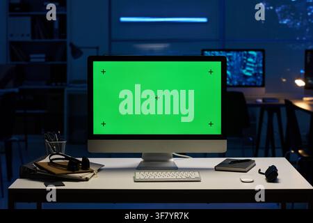 Computer Monitor with Green Screen on Desk in Dark Office Stock Photo
