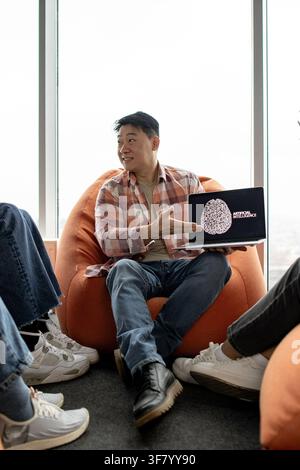 A man presents artificial intelligence concepts on a laptop while sitting on an orange beanbag chair with others around him Stock Photo