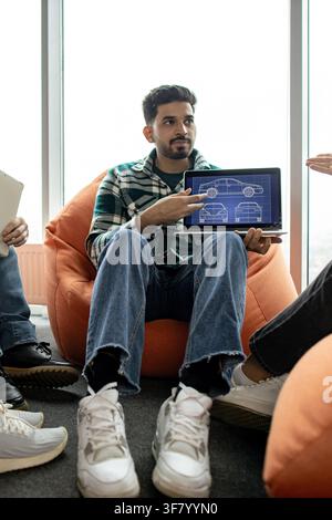 A young man in a casual outfit is presenting a car blueprint on his laptop to colleagues in a relaxed office setting Stock Photo