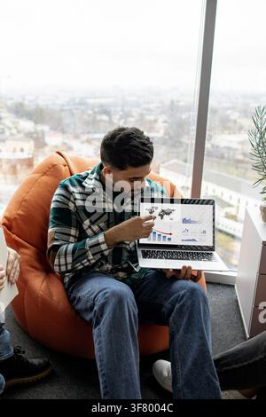 A man in a plaid shirt points to a laptop screen displaying world map and financial charts with a downward trend Stock Photo