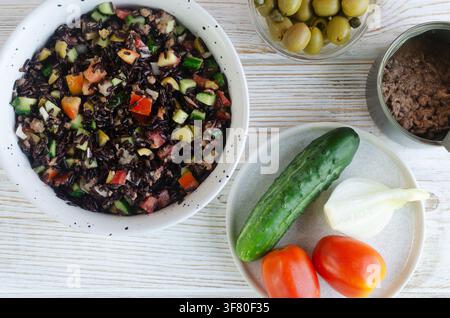 Camargue salad: black rice, can of tuna in oil, cucumbers, tomatoes, onions, olives and capers in a deep ceramic bowl on a white wooden background. Fr Stock Photo