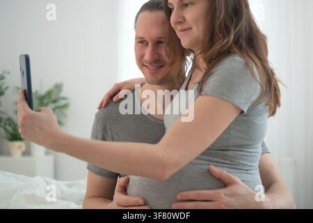 Happy pregnant woman and husband taking joyful selfie announcing pregnancy in bright bedroom. Expecting mother smiling at camera while partner poses with arm around shoulder excited about baby. Stock Photo