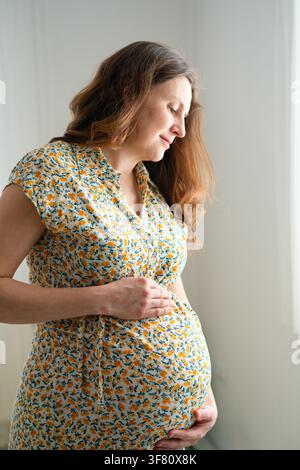 A pregnant woman in a floral dress stands enjoying the moment and her body indoors. The mother holding her belly shows serene, joyful self love and calm maternal contentment in a bright interior. Stock Photo