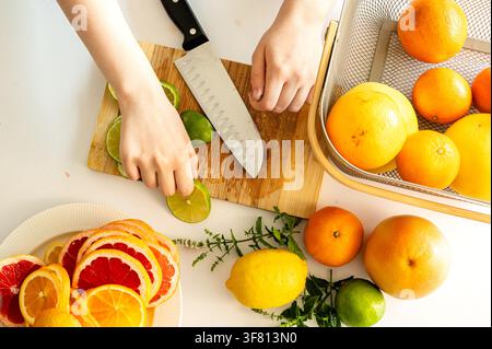 Overhead view of hands slicing a fresh lime on a wooden cutting board with a knife, surrounded by colourful citrus fruit and herbs, preparing ingredients for a healthy homemade summer lemonade. Stock Photo