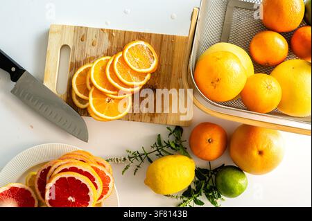 Slices of fresh orange arranged on a wooden cutting board with a knife, whole citrus and green herbs on a white table, preparing bright ingredients for a refreshing homemade citrus lemonade. Stock Photo
