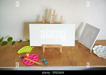 Blank canvas on an easel surrounded by paint palettes and brushes on a wooden table, creating a clean creative workspace with copy space. Stock Photo