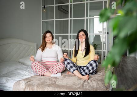 Two women practicing mindfulness meditation together at home sitting cross-legged on a bed. Wellness mental health routine and relaxation meditation p Stock Photo