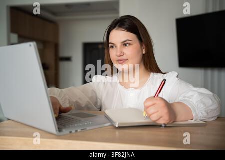 Young woman using laptop and taking notes at home to enhance learning experience. Remote work productivity and online education concept. Stock Photo