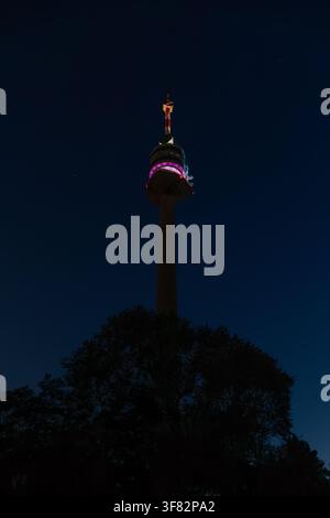 Low angle night view of illuminated Donauturm tower behind dark tree silhouette in Vienna Stock Photo