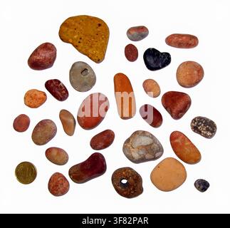 A variety of smooth reddish brown sea pebbles and natural hag stones are arranged with a cent coin for scale. The scene is set against a plain white b Stock Photo