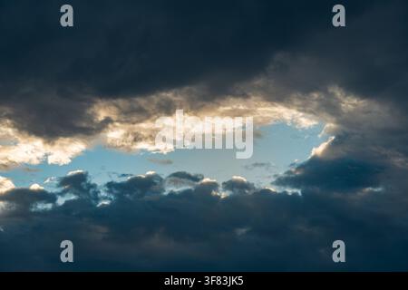 Dramatic dark storm clouds opening up to reveal a patch of clear blue sky illuminated by warm golden sunlight during late afternoon Stock Photo