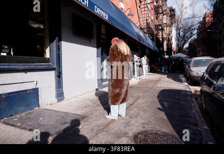 Woman in a fur coat waits outside a brunch spot on a sunlit New York City sidewalk Stock Photo