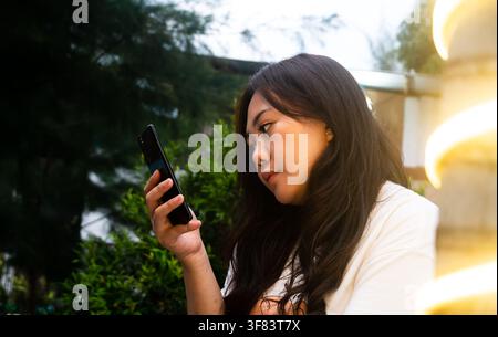 Side profile of a young Asian woman looking intently at her smartphone screen outdoors in the evening. Stock Photo
