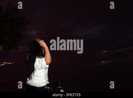 Rear view of a young woman with long dark hair adjusting her hair while standing outdoors at night against a dark twilight sky, casual lifestyle photo Stock Photo