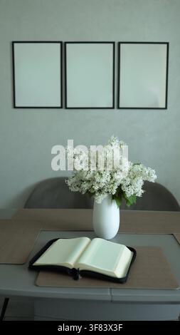Open blank book and white lilac bouquet in vase on wooden table with three empty picture frames on wall, interior mockup. Stock Photo