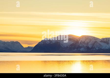Golden light spreads over snow-covered peaks and a calm fjord during dusk in Greenland with the sun setting behind mountains Stock Photo