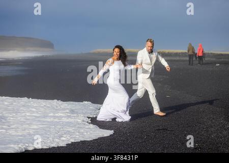 Newlywed couple races against waves on black sand beach in Iceland under moody skies Stock Photo