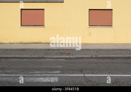 Yellow plaster house facade with two closed brown shutters. Sidewalk,street and weathered crosswalk marking in front. Background for copy space Stock Photo