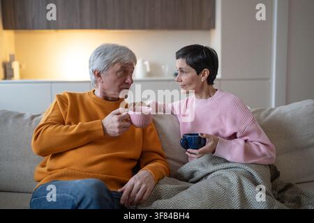 Senior husband and wife sitting on couch at home together drinking tea and talking with each other. Stock Photo