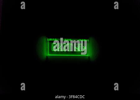 Glowing green exit sign with directional arrow on black background, symbolizing emergency escape and safety in public or commercial spaces. Stock Photo