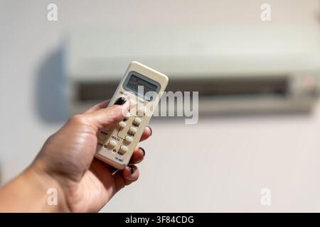Close-up of a hand using a remote control to operate a wall-mounted air conditioner in a modern indoor setting. Stock Photo