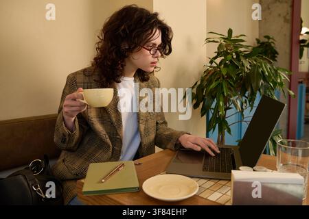 A young woman with curly hair types on her laptop, sipping coffee in a lively cafe Stock Photo