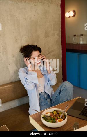 A young woman with curly hair happily works remotely at a cafe while having phone call Stock Photo