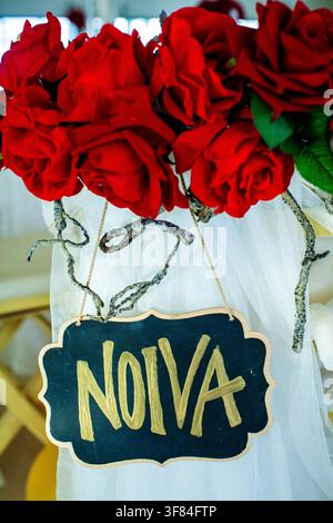 A wedding chair is decorated with red roses, dried branches, and a hanging chalkboard sign reading "Noivo". Stock Photo