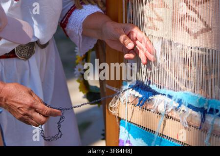 Woman dressed in traditional clothing works on a loom, creating a textile piece with vibrant colors and patterns, surrounded by floral elements Stock Photo