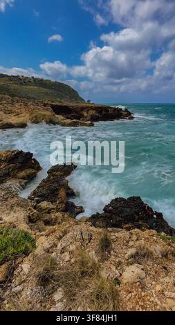 Vertical view of stormy turquoise Mediterranean sea waves crashing against rocky coastline and cliffs under blue sky with clouds in Crete island Greec Stock Photo