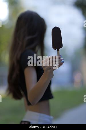 Young woman holding a melting chocolate ice cream bar on a stick, enjoying a refreshing sweet treat outdoors in warm weather, symbolizing summer leisu Stock Photo