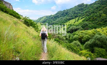 A wide shot of a blonde woman hiking on a narrow dirt trail along a steep, grassy hillside. She is viewed from behind, wearing a grey t-shirt, light c Stock Photo