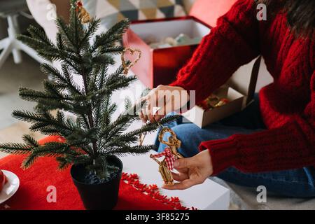 Woman hands hang straw ornament on branch of small potted tree on table. Handmade rustic christmas ornaments, organic tree decoration style, zero Stock Photo