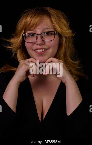 Young woman with blonde hair smiling and resting chin on hands against black background Stock Photo