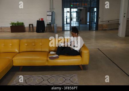 Woman working with a notebook on a bright yellow leather sofa in an open plan lounge. Stock Photo