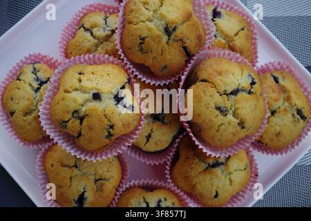 Golden-brown muffins with visible blueberries sit in pink paper liners, arranged on a white plate. Their tops are rounded and slightly cracked, showin Stock Photo