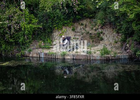 Vinkovci, Croatia - May 09.2026:A solitary angler sits on a folding chair at a small riverside bank, managing multiple fishing rods beside calm reflec Stock Photo