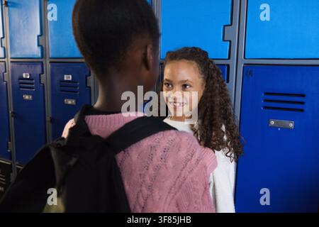 African American teen females chatting at school by blue lockers with black backpack, pink sweater. Hallway, friends, casual, youth, talking, interact Stock Photo